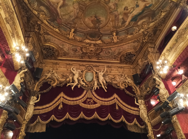 Theaterinnenraum mit roter Gardine, Kronleuchtern, Deckenplastiken, Lichtern und einem Gemälde, das eine Bühne und einige Personen am Boden zeigt.