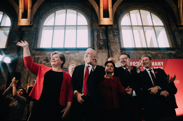 Eine Gruppe von Menschen, wahrscheinlich Labour-Parteimitglieder, die vor einem Publikum applaudieren, mit einem Banner und beleuchteten Fenstern im Hintergrund.