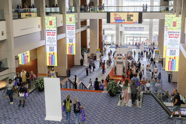 Eine große Gruppe von Menschen, die durch ein großes Gebäude mit Bannern, Topfpflanzen, Rolltreppen, Geländern, Säulen, Texttafeln, Glastüren und anderen Gegenständen geht, einige tragen Taschen, wahrscheinlich an der Nationalen Buchmesse teilnehmend.