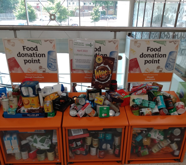 Orange Schübe mit Lebensmittelspenden, darunter Flaschen, Dosen und Kartons, mit informativen Schildern an der Wand, durch Glasfenster betrachtet, die Außerfahrzeuge, Bäume, Gebäude und Himmel zeigen.