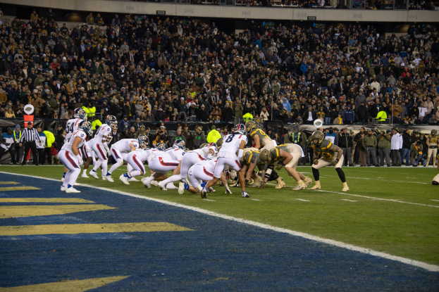 Ein Fussballspiel mit Spielern in Helmen auf dem Feld, Zuschauern, Geländern und Tafeln mit Text im Hintergrund.