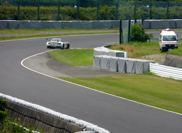 Ein Rennwagen fährt eine Strecke entlang mit einem Polizeiwagen im Hintergrund, umgeben von Gras, Pflanzen, Bäumen und Zaun, mit einem Fahrzeug auf der rechten Seite und Text unten.