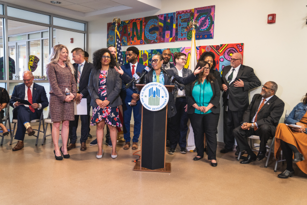 Eine Gruppe von Menschen, die vor einem Podium mit Mikrofon und einer Tafel mit der Aufschrift "Sancho-Stadtrat tritt dem Stadtrat bei" stehen und sitzen, mit Flaggen, Feuerlöscher und Deckenleuchten an der Rückwand.