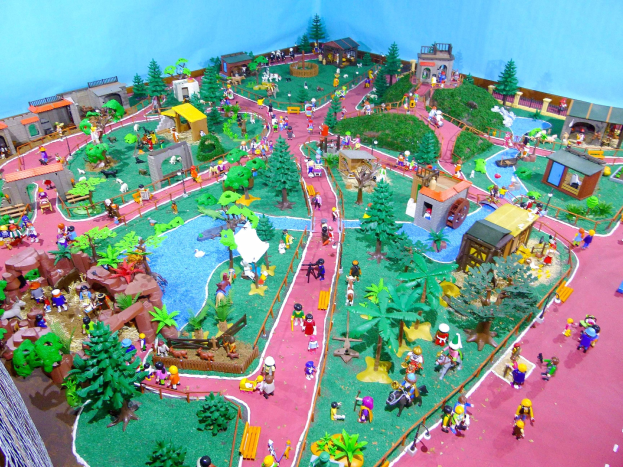 Ein Miniatur-Zoo-Modell mit zahlreichen Besuchern, umgeben von Bäumen, Häusern, Pflanzen, Gras und einem Zaun, vor einer Hintergrundwand mit Text unten.